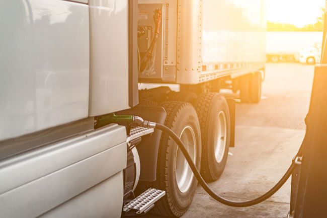 A semi truck is seen at a fueling station with the gas hose in the tank and a sunburst in the background.
